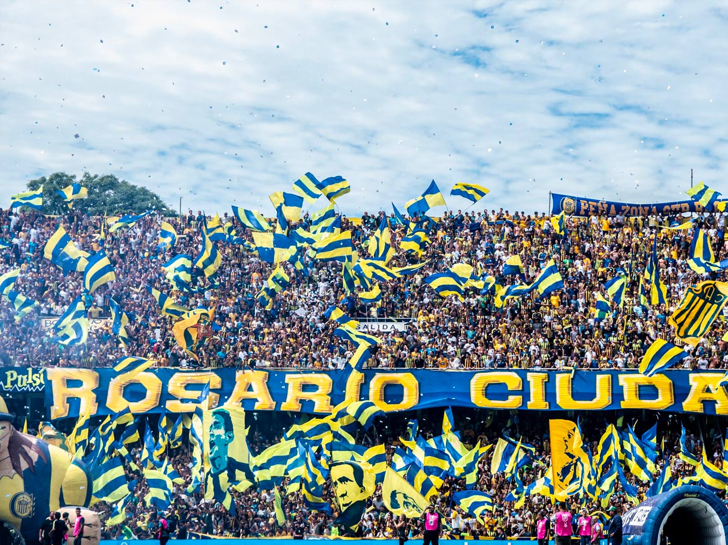 Hinchada canalla desplegando una bandera gigante con la leyenda 'Rosario Ciudad' entre banderas y papel picado
